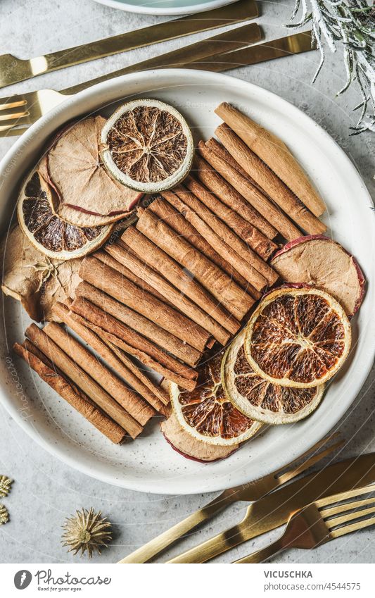 Wintergewürze, getrocknete Äpfel und Orangen in weißer Schale auf grauem Küchentisch mit goldenem Besteck. Zimtstangen und Winterfrüchte für Glühwein und Weihnachtspunsch. Ansicht von oben.