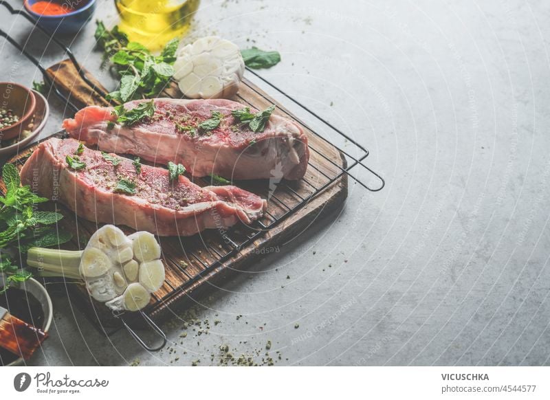 Rohes Fleisch auf Grillrost mit Geschmack Zutaten: Knoblauch, Kräuter und Gewürze auf grauem Beton Küchentisch. Kochen Vorbereitung mit Steak für bbq. Draufsicht mit Kopierbereich.