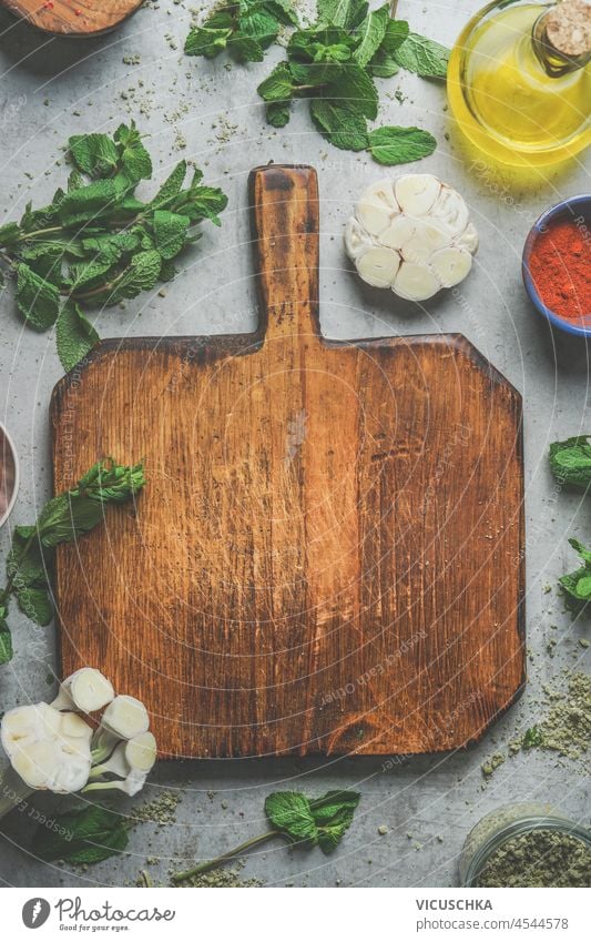 Lebensmittel Hintergrund mit leeren Holzschneidebrett, Kräuter, Knoblauch, Olivenöl, Gewürze auf grauem Beton Küchentisch. Kochen mit frischen, geschmackvollen Zutaten. Draufsicht mit Kopierbereich.