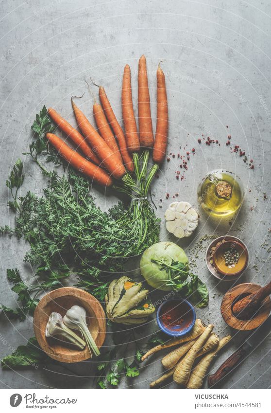 Verschiedene gesunde vegetarische Zutaten: Karotten, Pastinaken, Kürbis, Kohlrabi, Knoblauch, Gewürze und Küchenutensilien auf einem hellgrauen Betonküchentisch. Leckeres Kochen mit Wurzelgemüse. Ansicht von oben.