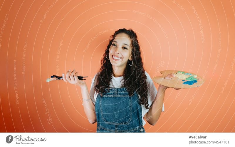 Junge Künstlerin hält eine Palette und einen Pinsel über isolierten orangefarbenen Hintergrund und schaut zur Seite. Afrikanische lockiges Haar Frau. Kunst und Kreativität Konzept, Kopie Raum. Lächeln