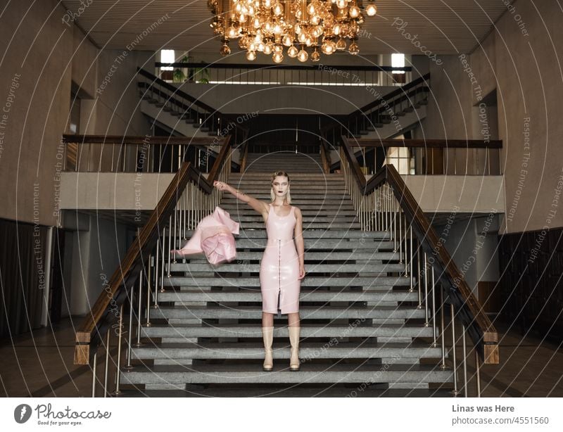 Ein altes Herrenhaus ist ein perfekter Ort für ein wunderschönes Model, um ein rosa Latex-Outfit und ein hübsches Gesicht zu zeigen. Massiver Kronleuchter, Retro-Treppe, rosa Outfit. Die wichtigsten Themen dieser Szene.