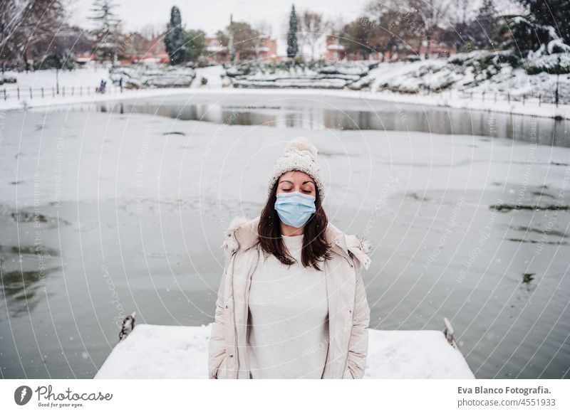 entspannte frau mit geschlossenen augen und gesichtsmaske auf dem pier vor einem zugefrorenen see in der stadt. reisen und lebensstil im winter in der stadt während des pandemischen coronavirus