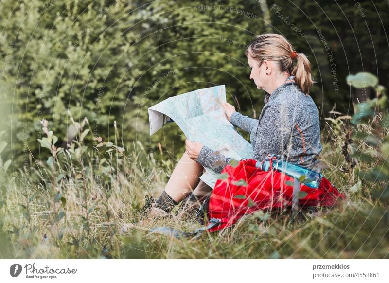 Frau mit Rucksack, die während eines Ausflugs in den Bergen eine Pause macht und auf eine Karte schaut, während sie im Gras sitzt Sommer aktiv Landkarte