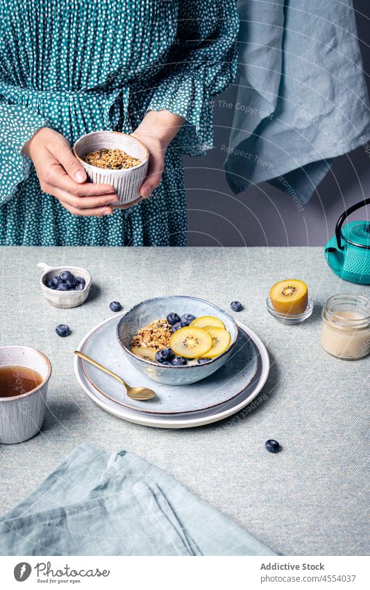 Frau gibt Müsli in eine Schüssel mit Obst hinzufügen Blaubeeren Kiwi Lebensmittel Frühstück Ernährung Portion Morgen Tee selbstgemacht Bestandteil