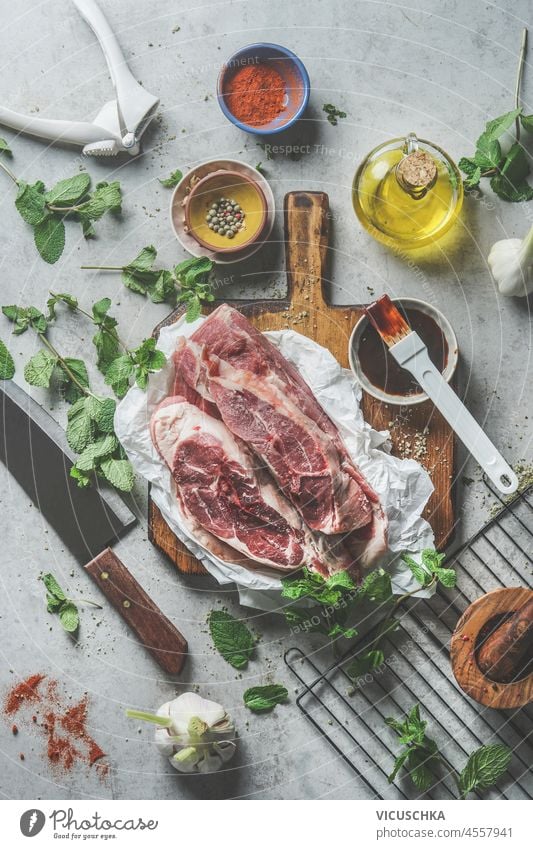 Rohe Fleischstücke auf Holzschneidebrett mit Öl, Gewürzen, Marinade, Kräutern und Kochutensilien auf grauem Küchentisch. Kochen und Grillen Vorbereitung zu Hause mit geschmackvollen hausgemachten Marinade. Ansicht von oben.