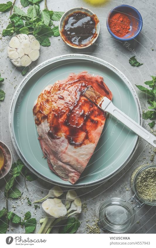 Rohes Fleisch mit hausgemachter Marinade auf blauem Teller mit Knoblauch, Gewürzen und Kräutern auf grauem Betonhintergrund. Fleischzubereitung für BBQ oder Grill. Ansicht von oben.