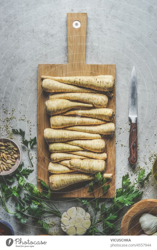 Rohes Pastinakenwurzelgemüse auf Holzschneidebrett mit Küchenmesser, Kardamon, Knoblauch und Kräutern auf grauem Betonküchentisch. Zubereitung von saisonalem Herbstgemüse zu Hause. Ansicht von oben.