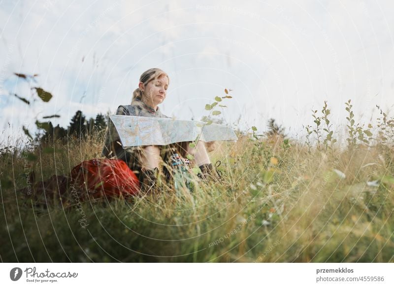 Frau mit Rucksack, die während eines Ausflugs in den Bergen eine Pause macht und auf eine Karte schaut, während sie im Gras sitzt Sommer aktiv Landkarte