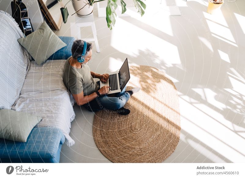 Älterer Mann mit Kopfhörern und Laptop im Wohnzimmer sitzend benutzend Loft zuhören Innenbereich Stil Liege Sofa Kälte ruhen Zeitvertreib Hobby unterhalten