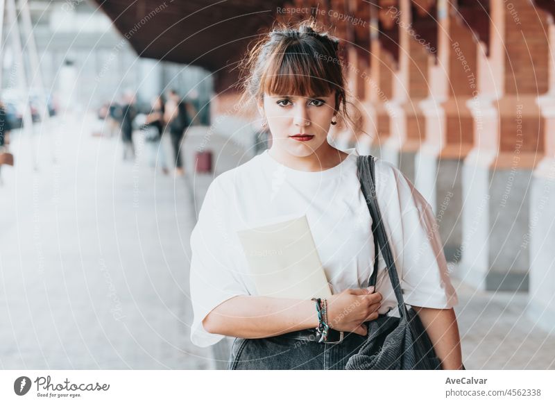 Junge schöne rote Kopf schaut ernst in die Kamera Hipster Frau.Halten Sie ein Buch in der Nähe des Bahnhofs. Trendy Mädchen im Sommer-T-Shirt.nachdenklich weiblich posiert auf der Straße in der Nähe einer Mauer.