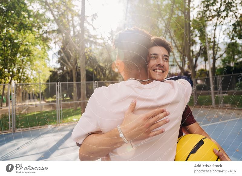 Positiv gestimmte Sportler mit Basketball, die sich auf einem Sportplatz umarmen Männer Ball Freund Spieler Zusammensein Umarmung Teenager Klassenkamerad