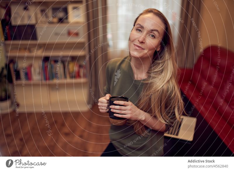 Fröhliche Frau mit Kaffee im Zimmer Heißgetränk Wohnzimmer heimisch Appartement Liege Laptop Getränk heimwärts Sofa positiv Netbook Dame Licht Tasse Inhalt