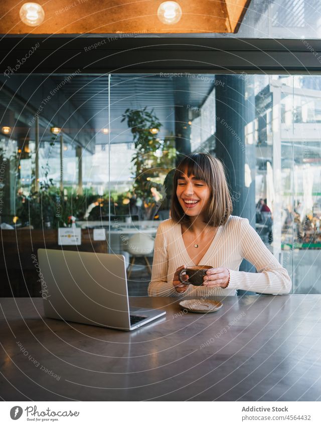 Fröhliche Frau mit einer Tasse Kaffee und Blick auf einen Laptop Videoanruf Café benutzend Browsen Gerät Anschluss abgelegen Gespräch freiberuflich Telefon