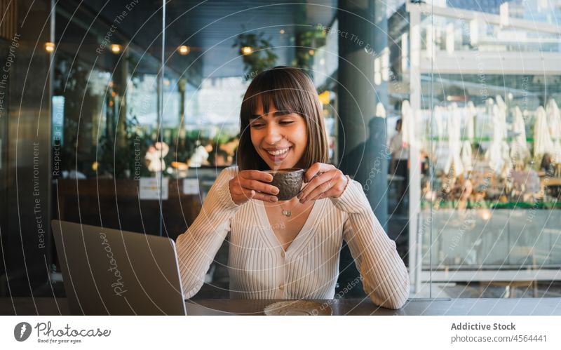 Fröhliche Frau mit einer Tasse Kaffee und Blick auf einen Laptop Videoanruf Café benutzend Browsen Gerät Anschluss abgelegen Gespräch freiberuflich Telefon