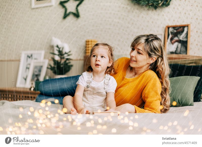 Mutter und Tochter lesen ein Buch und lächeln, sie sitzen zu Hause auf dem Bett. Neujahr, Weihnachten, Familie, gemeinsame Zeit Mama Weihnachtsbeleuchtung