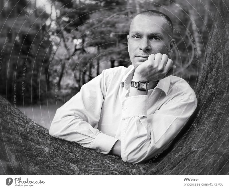 Der junge Mann lehnt an einem Baum, stützt den Kopf auf und schaut freundlich und abwartend Portrait Natur Außenaufnahme zuhören aufmerksam nett attraktiv Uhr