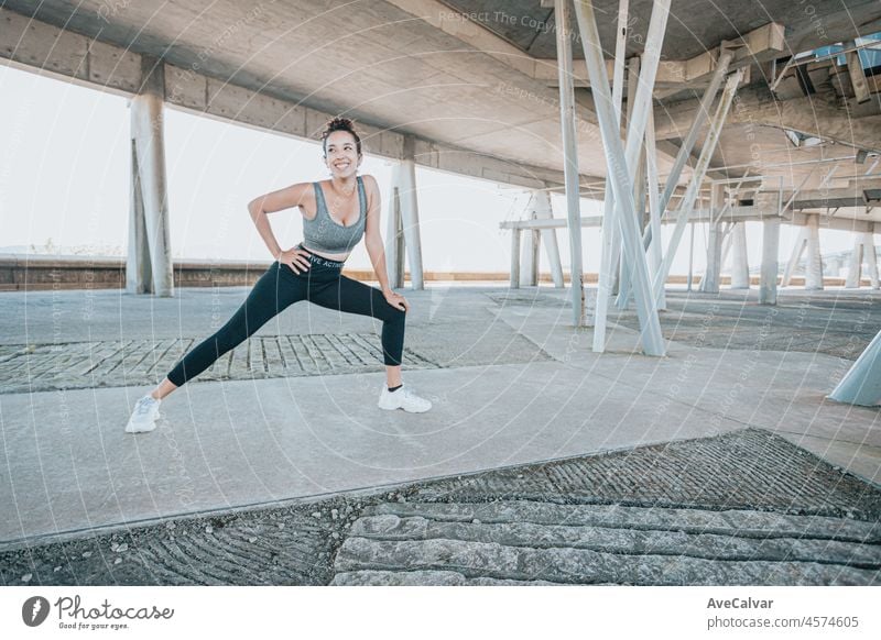 Lächelnde junge afrikanische Frau übt Yoga, streckt die Beine über die Matte, Gesundheitslehrer. Trainiert, trägt Sportkleidung, schwarze Hose und Oberteil, städtische Outdoor-Stadt in voller Länge, Blick in die Kamera