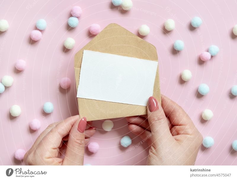 Hände mit Blankokarte und Umschlag in der Nähe von pastellfarbenen Pom-Poms auf rosa Ansicht von oben Hochzeit Postkarte Kuvert Pompom Attrappe Draufsicht