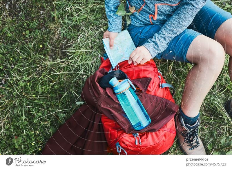 Frau mit Rucksack, die während eines Ausflugs in den Bergen eine Pause einlegt und eine Karte herausnimmt, während sie im Gras sitzt Sommer aktiv Landkarte