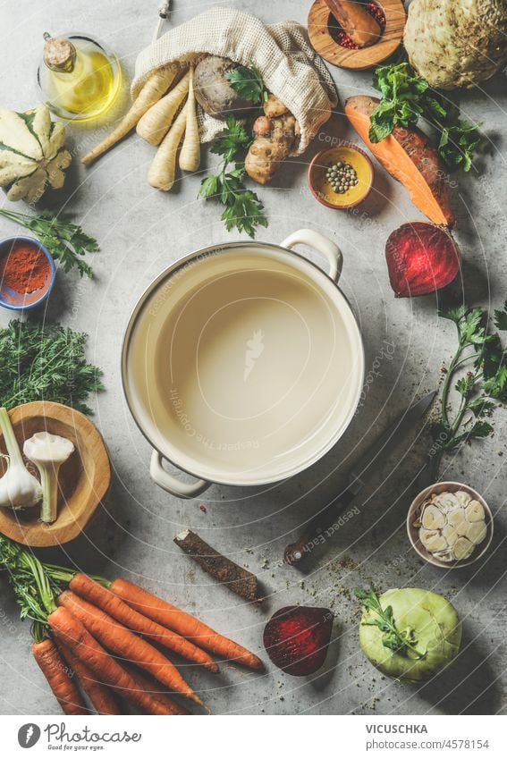 Verschiedene Gemüse um leeren Kochtopf: Karotten, Kohlrabi, Rote Bete, Pastinake, Knoblauch, Süßkartoffel, Kräuter und Gewürze auf grauem Betonküchentisch. Kochen gesundes Essen zu Hause. Ansicht von oben.