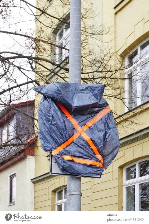 mit einer blauen Plastiktüte und orangefarbenem Klebestreifen verhülltes Verkehrsschild inklusive Zusatzschild am Straßenrand vor einem Wohnhaus / Baumaßnahme / Straßenverkehr