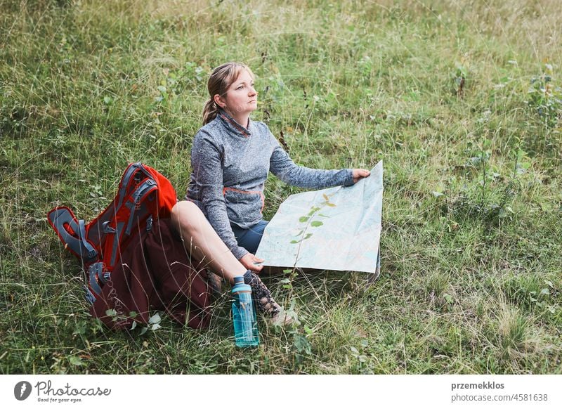 Frau mit Rucksack, die während eines Ausflugs in den Bergen eine Pause macht und auf eine Karte schaut, während sie im Gras sitzt Sommer aktiv Landkarte