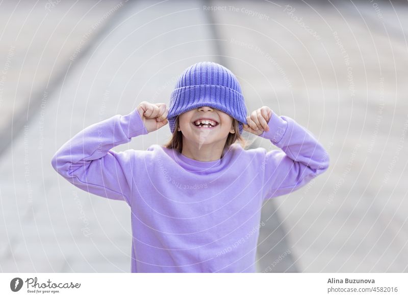 Nette kleine kaukasische Mädchen acht Jahre alt mit blonden Haaren lächelnd im Freien. Kind trägt stilvolle Shirt und Strickmütze violette Farbe. Trendy Farbe des Jahres 2022 sehr peri