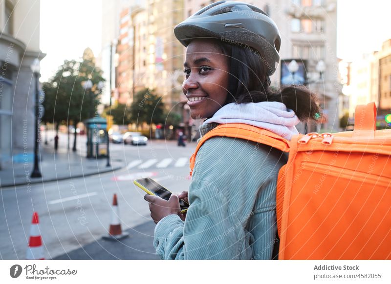 Schwarze Lieferfrau durchsucht Smartphone in der Nähe der Straße Frau Versand Rucksack Dienst App Orden prüfen Arbeit Fahrbahn Straßenrand Thermo Spaziergang