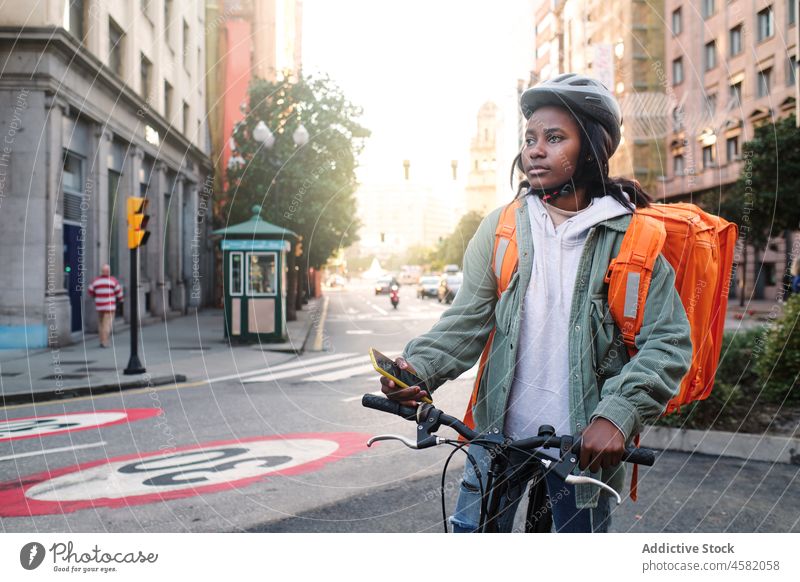 Schwarze Zustellerin auf dem Fahrrad Frau Versand Rucksack App Dienst Smartphone Orden Straße Reiter Mitfahrgelegenheit Fahrbahn Arbeit Thermo Gebäude Großstadt