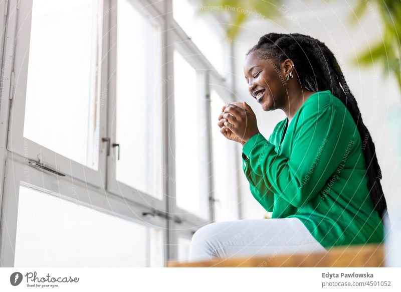 Junge Frau mit einer Tasse Kaffee und Blick aus dem Fenster schwarz Jahrtausende Hipster im Innenbereich Loft natürlich Erwachsener attraktiv gelungen Menschen