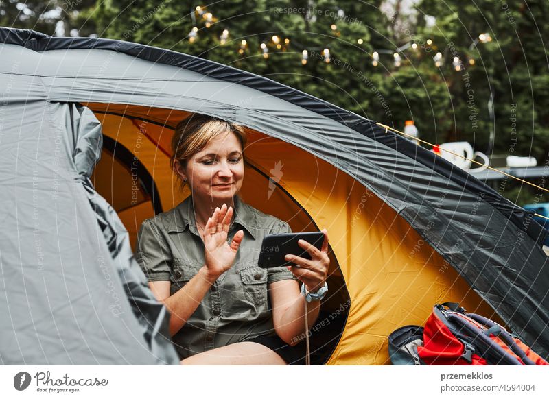 Eine Frau führt einen Videoanruf mit Freunden über ihr Smartphone, während sie im Zelt beim Camping sitzt. Frau entspannt im Zelt während des Sommerurlaubs