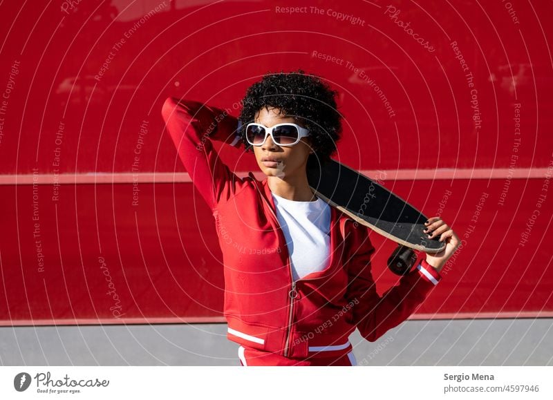 Afroamerikanische Frau mit kurzen Haaren, Sonnenbrille und roter Sportkleidung mit ihrem Skateboard Afroamerikaner Spanien Kleidung Wand Hintergrund Stehen