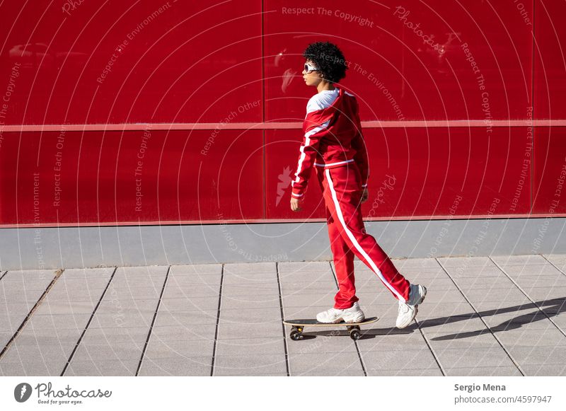 Afroamerikanische Frau mit kurzen Haaren und roter Sportkleidung fährt auf ihrem Skateboard mit rotem Hintergrund Afroamerikaner Spanien Kleidung Wand Stehen