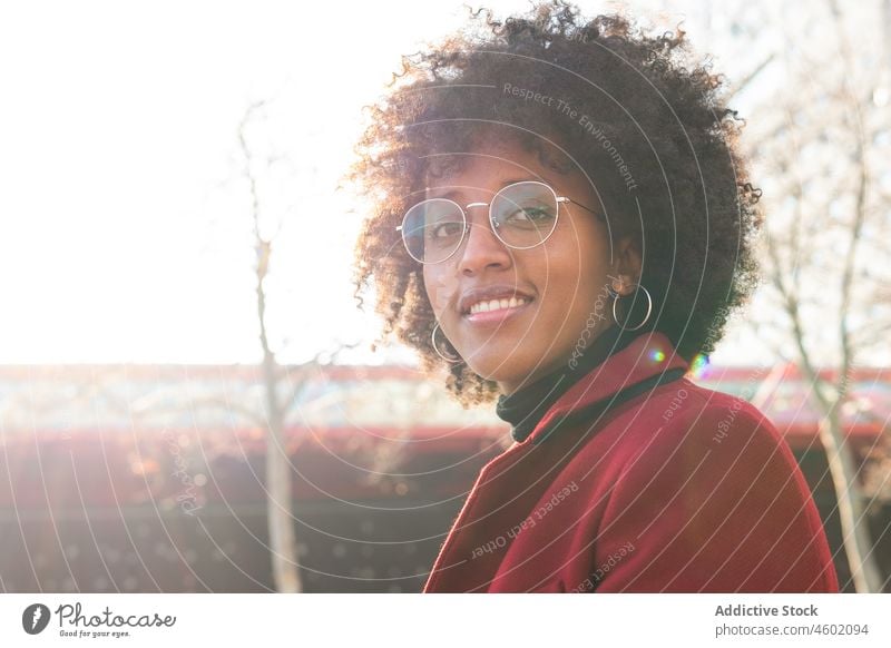 Lächelnde afroamerikanische Frau in karmesinrotem Mantel und mit Augengläsern Afro-Look Frisur positiv krause Haare Brille lässig Porträt heiter Sonnenlicht