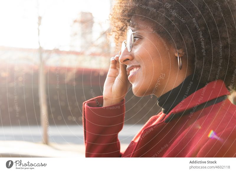 Lächelnde afroamerikanische Frau in karmesinrotem Mantel und mit Augengläsern Afro-Look Frisur positiv krause Haare Brille lässig Porträt heiter Sonnenlicht