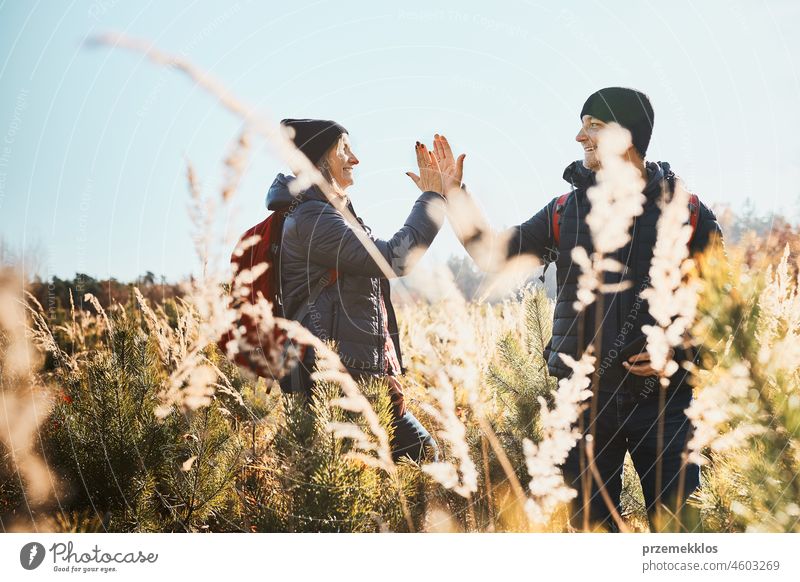 Freunde, die einen Ausflug in die Natur genießen, geben High Five während des Urlaubstages. Wanderer mit Rucksäcken auf dem Weg in die Berge. Wandern durch hohes Gras entlang des Weges in der Wiese an einem sonnigen Tag