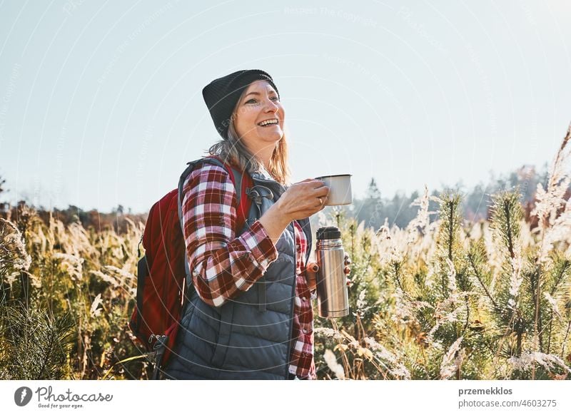 Lächelnde Frau, die sich entspannt und den Kaffee während einer Urlaubsreise genießt. Frau, die auf einem Wanderweg steht und wegschaut, hält eine Tasse Kaffee und eine Thermoskanne. Frau mit Rucksack Wandern durch hohes Gras entlang Weg in den Bergen