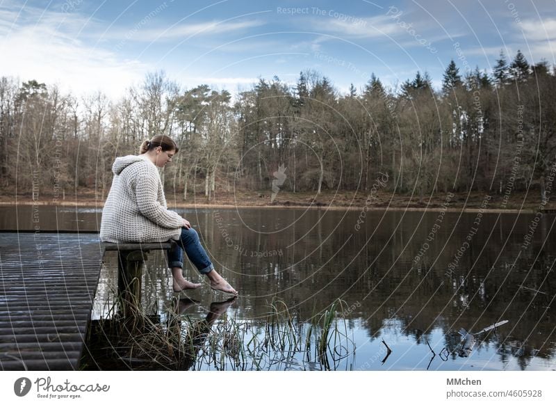 Frau sitzend am Steg, streckt den Fuß ins Wasser See Kratersee barfuß Wasseroberfläche blau kalt Spiegelung Natur Wald Landschaft Teich windstill Farbfoto