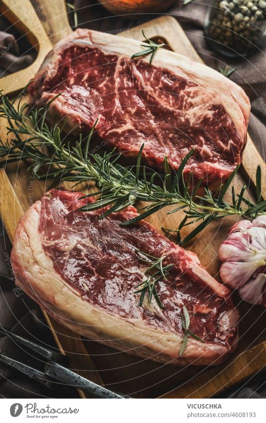 Nahaufnahme von zwei roh marmorierten Steaks mit frischem Rosmarin und Knoblauch auf einem rustikalen Schneidebrett. Ansicht von oben abschließen Draufsicht