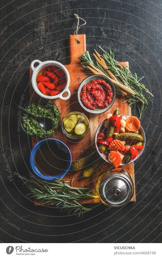 Schalen mit verschiedenen eingelegten Gemüsesorten auf einem hölzernen Schneidebrett mit frischen Kräutern, Pesto und hausgemachtem Ketchup auf dunklem, rustikalem Hintergrund. Ansicht von oben