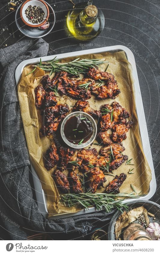 Gebratene Hähnchenflügel mit BBQ-Sauce auf Backblech auf dunklem Küchentisch mit Zutaten, Ansicht von oben Lebensmittel gebraten Abdeckfolie dunkel Tisch