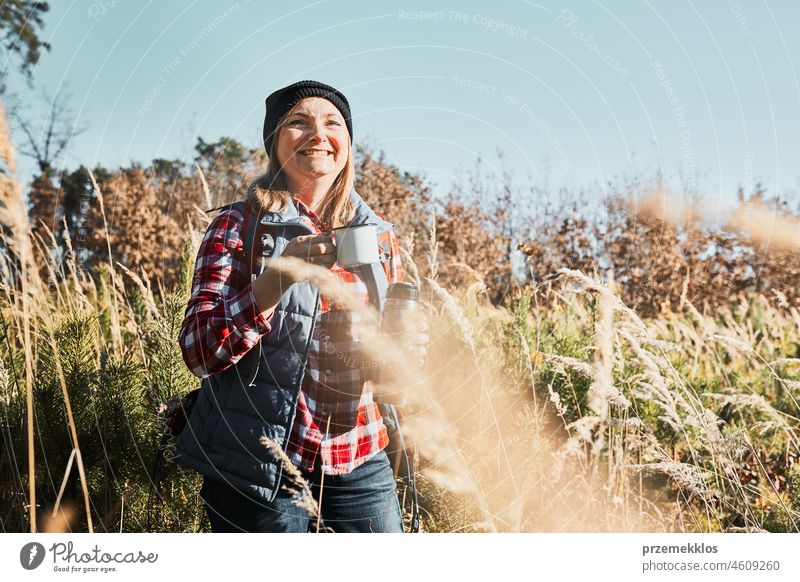 Lächelnde Frau entspannt sich und genießt den Kaffee während einer Sommerreise. Frau, die auf einem Wanderweg steht und wegschaut und eine Tasse Kaffee hält. Frau mit Rucksack Wandern durch hohes Gras entlang Weg in den Bergen