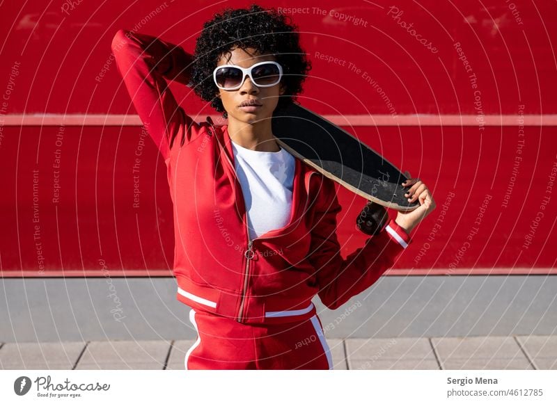 Afroamerikanische Frau mit kurzen Haaren, Sonnenbrille und roter Sportkleidung mit ihrem Skateboard Afroamerikaner jung Junge Frau Erwachsener erwachsene Frau