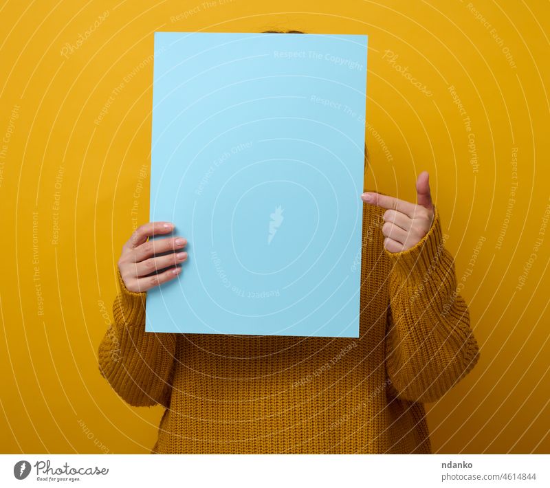 Frau im orangefarbenen Pullover hält ein leeres Blatt Papier auf gelbem Hintergrund. Platz für eine Inschrift, Werbung, Information blanko Person Schot Plakat