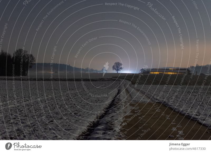 Feld und Weg mit Schnee am Abend mit landwirtschaftlichem beleuchtetem Betrieb im Hintergrund Winter Feldweg" Landwirtschaft landwirtschaftliche Fläche