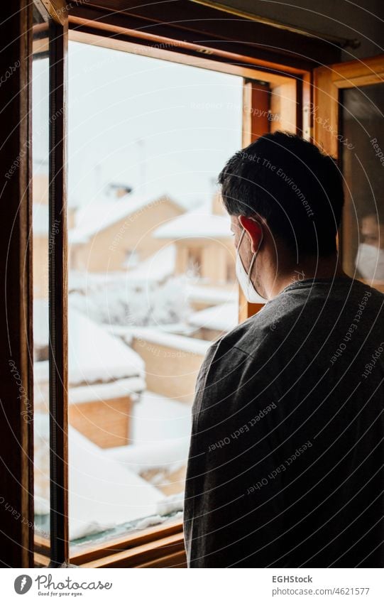 Junger Mann mit Maske in Quarantäne, der durch das Fenster seines Hauses den Schneefall betrachtet Blick Männer jung begrenzt Einsperrung Mundschutz