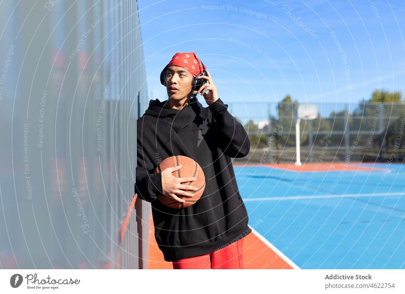 Asiatischer Mann mit Basketball, der auf einem Spielplatz Musik hört Kopfhörer Gesang Porträt Ball Sport Hobby Training asiatisch Zaun Sportpark männlich