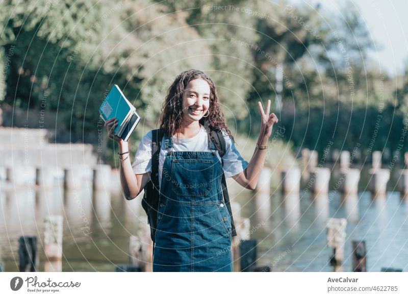 Junge arabische afrikanische Studentin hält Bücher und tut das V-Zeichen an der Universität im Freien mit Positivität Haltung glücklich, Sonnenuntergang ambient. Neue Studiengang, Selbstverbesserung und Lernen beim Lesen.