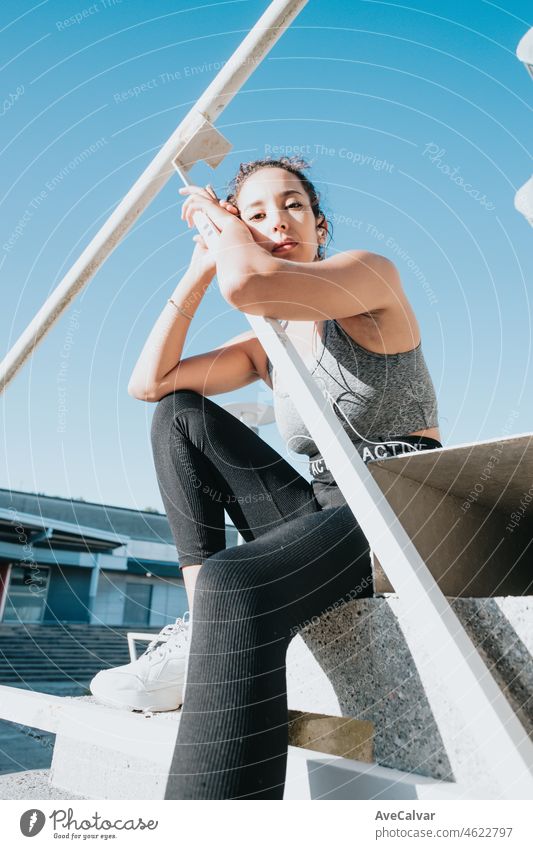 Junge arabische afrikanische Frau auf Ausbildung Sport tragen Leggins und Top sitzen auf der Treppe beim Hören von Musik ruht nach dem Training. Serious Blick auf Kamera, Schönheit jungen Athleten städtischen Stadt Hintergrund